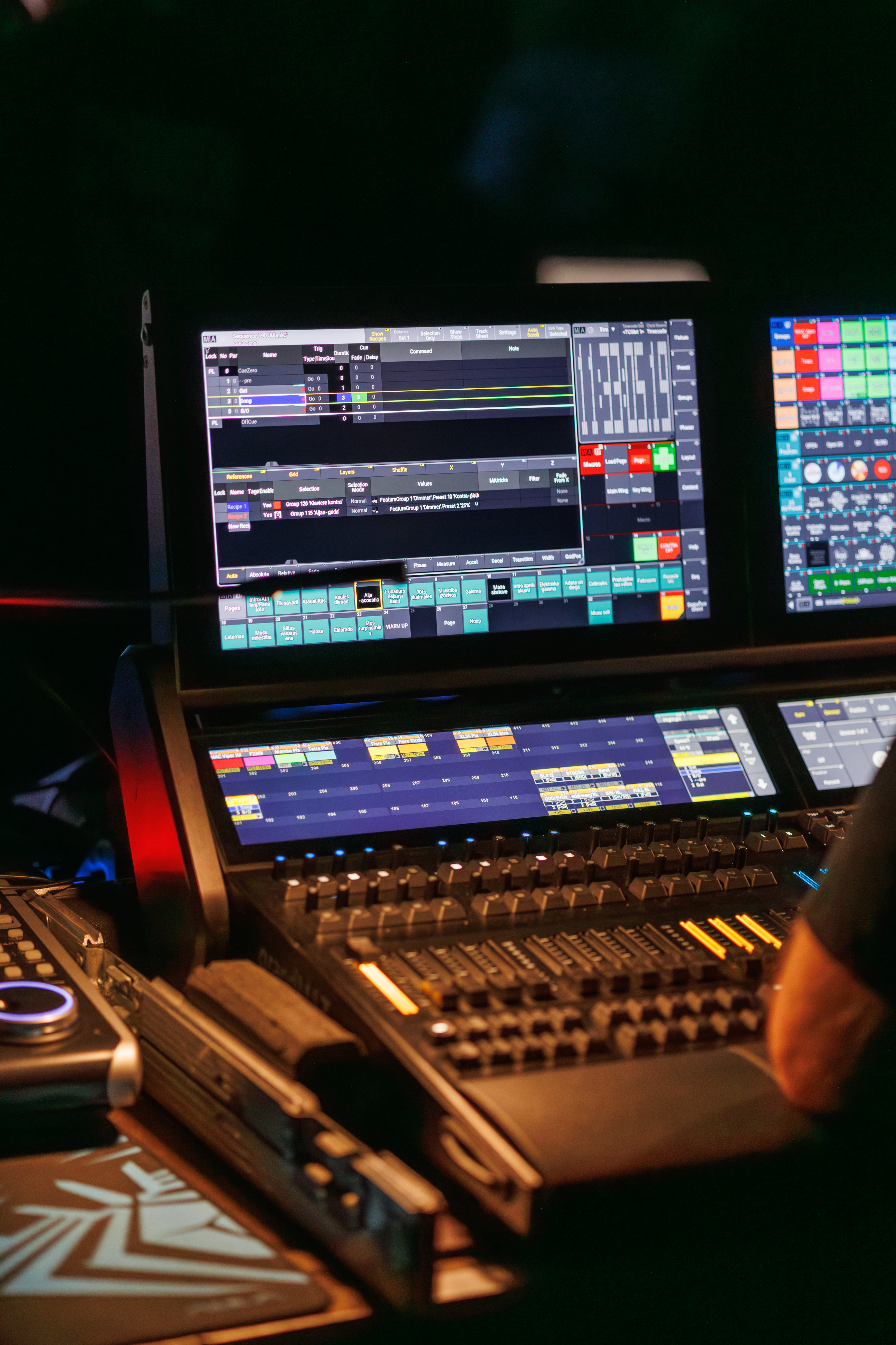 Technicians operate lighting and audio control consoles during a live event, with illuminated screens and a blurred crowd in the background, creating a dynamic concert production atmosphere